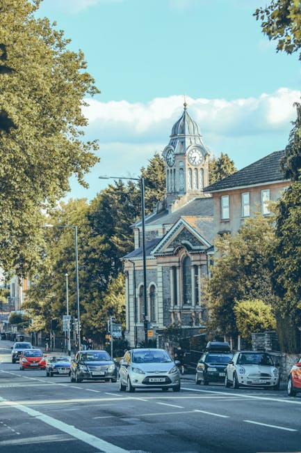 A street scene in Bow featuring several parked and moving cars alongside a sidewalk lined with large leafy trees. In the background, there is a historic building with a prominent clock tower, ornate architectural details, and a steeply pitched roof. The street appears to be part of a home relocation route, with visible space for loading and unloading near the curb. The image is well-lit with natural daylight, and the environment reflects a typical urban setting suitable for house removals and furniture transport, as managed by companies like bowremovals.org.uk, which specializes in house removals and moving services.