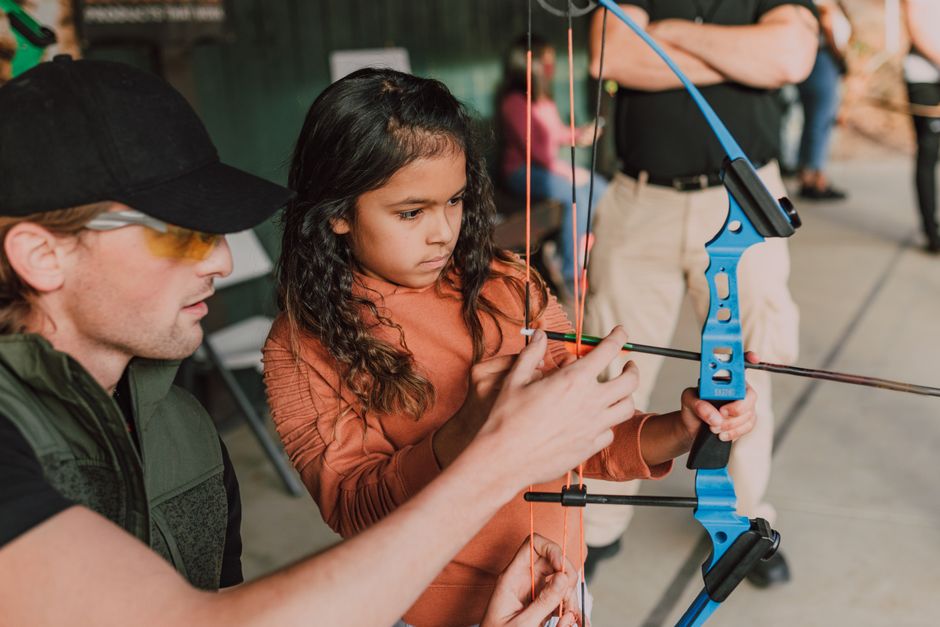 A young girl with long, dark, curly hair wearing an orange long-sleeved top is holding a blue and black compound bow with her left hand, while an adult male, wearing a black cap, yellow-tinted glasses, and a dark green vest, is assisting her in adjusting or aiming the bow. The scene takes place outdoors on a paved surface, with other people visible in the background. The girl appears focused, and the adult is guiding her hands during a target practice session or outdoor activity. The image captures the moment of learning or instruction related to archery, with equipment such as the bow and arrow present, reflecting a setting associated with outdoor recreation or a community event possibly connected to moving or packing activities managed by [COMPANY_NAME] for home relocation purposes.